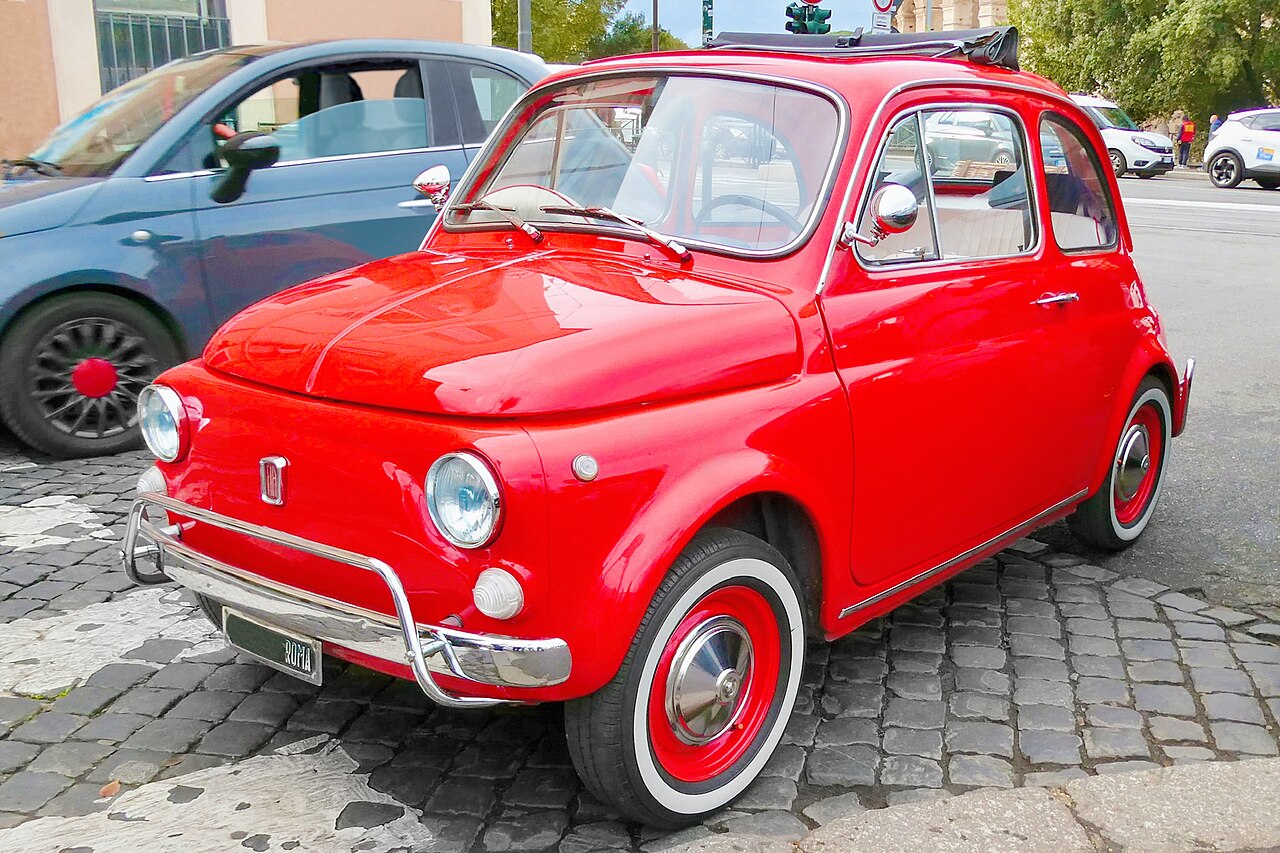 Petite Fiat 500 rouge classique garée sur des pavés en ville, vue avant-gauche.
