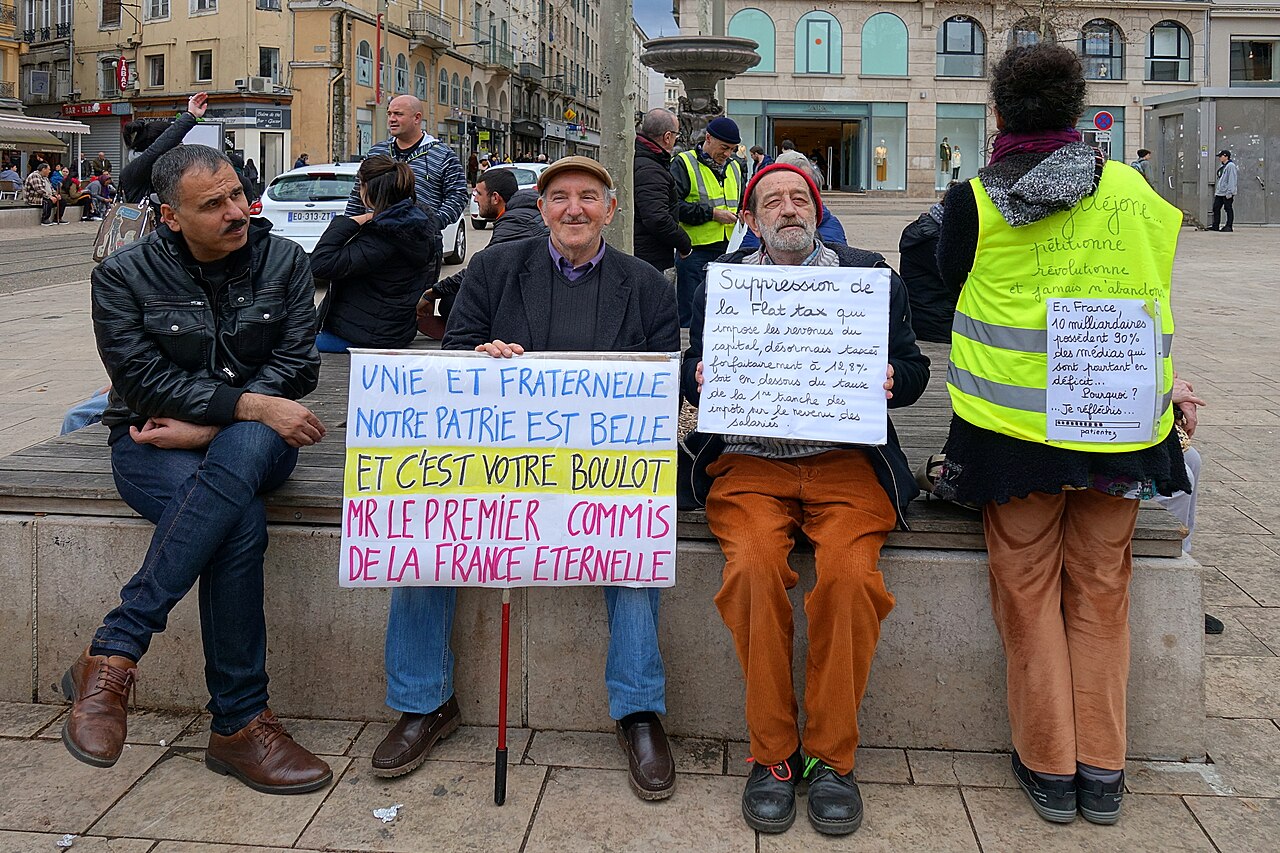 Plusieurs manifestants portant des gilets jaunes et tenant des pancartes assis sur un muret en centre‑ville.