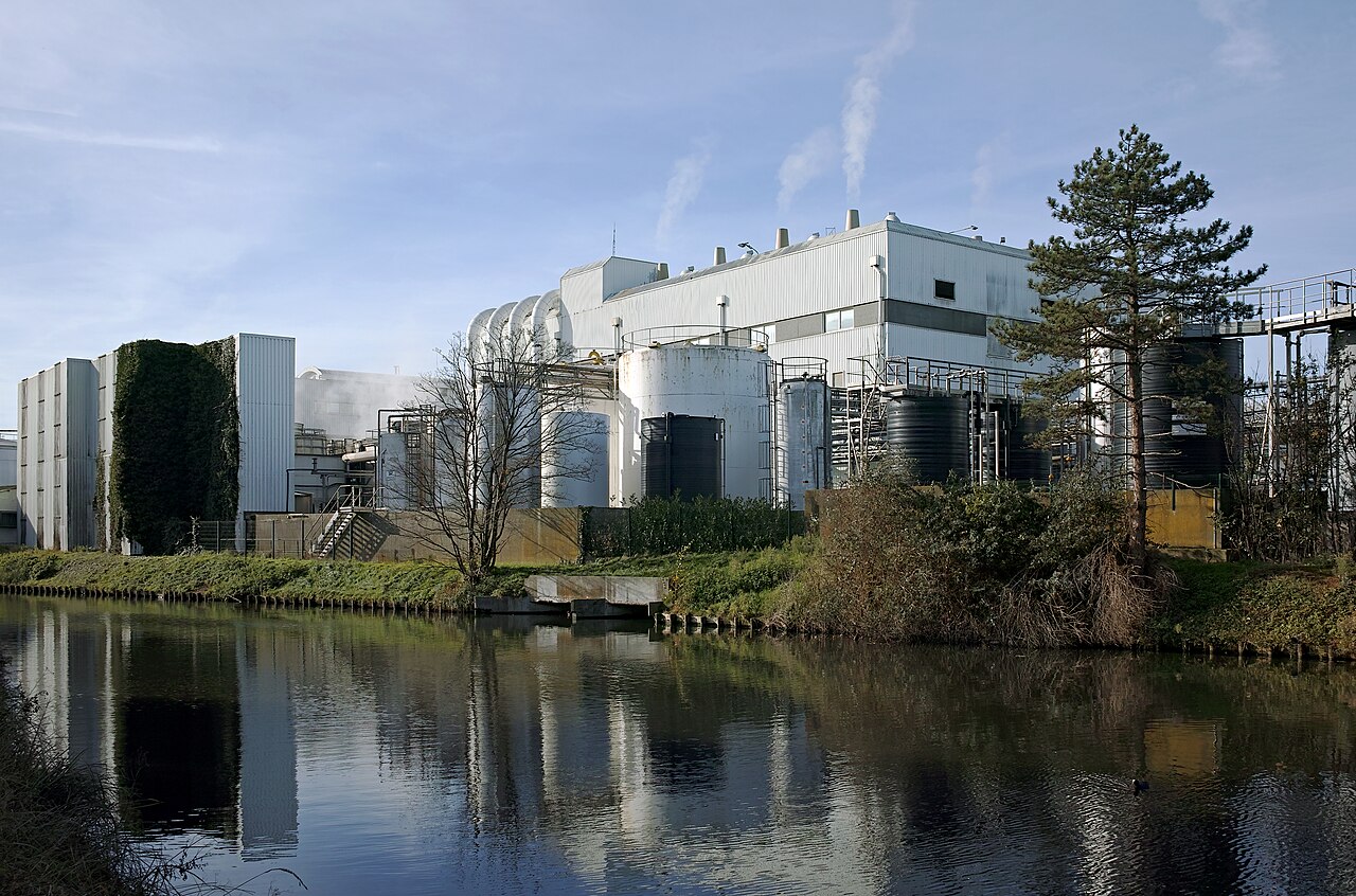 Façade et réservoirs d’une usine industrielle bordant un canal, avec fumées légères et reflets sur l’eau.