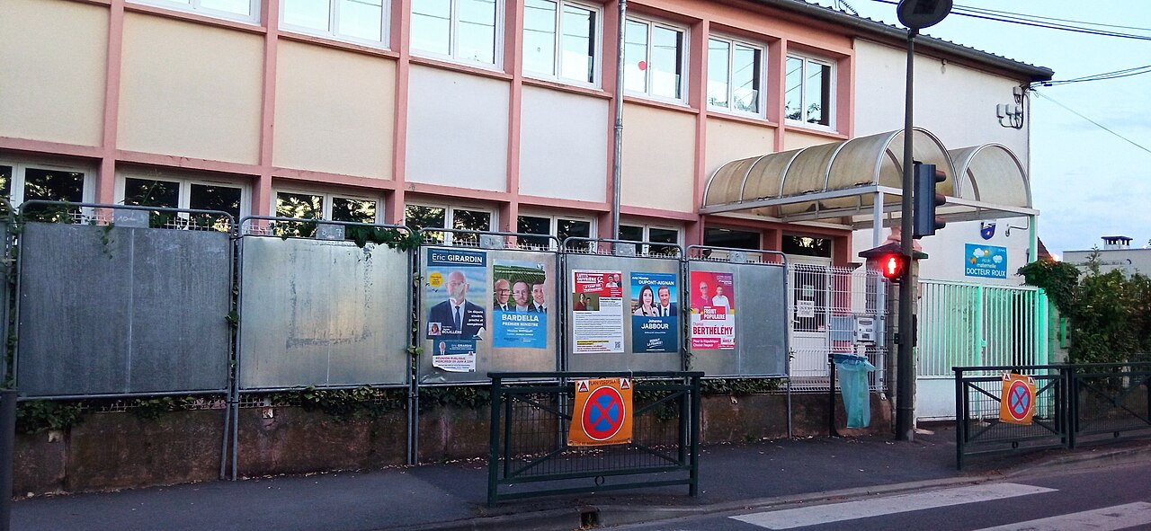 Panneau d'affichage électoral sur le trottoir devant la façade d'un bâtiment scolaire.