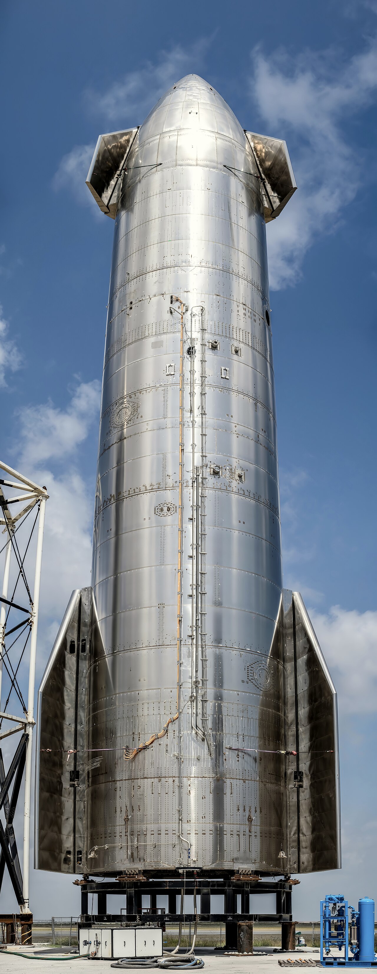 Vue verticale rapprochée d’un prototype Starship en métal brillant posé à côté d’une tour de lancement, sous un ciel bleu.